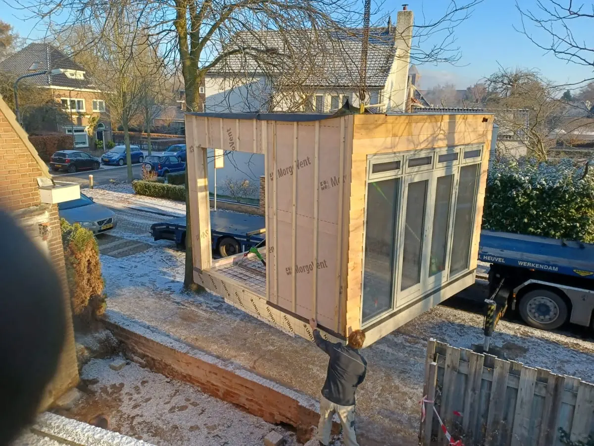Prefab aanbouw hangt aan de kraan boven de fundering naast de woning