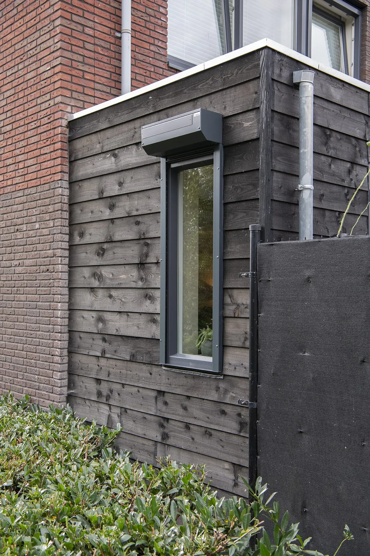 Zwarte houten uitbouw met smal raam naast de bakstenen gevel